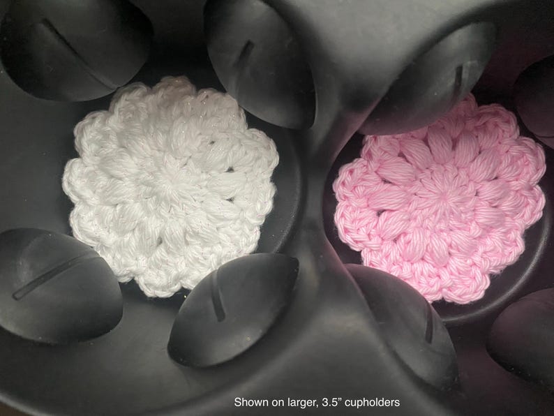 May include: Two circular crochet coasters, one white and one light pink, featuring a textured floral pattern. These coasters are positioned inside a black plastic cup holder assembly, with text at the bottom stating shown on larger, 3.5" cupholders.