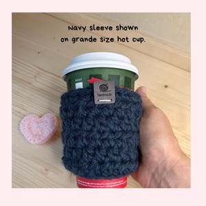 May include: A navy blue crocheted cup sleeve featuring a small brown rectangular tag with the text handmade and a yarn ball icon. The sleeve is wrapped around a green grande size hot cup with a white lid. Text reads: Navy sleeve shown on grande size hot cup.