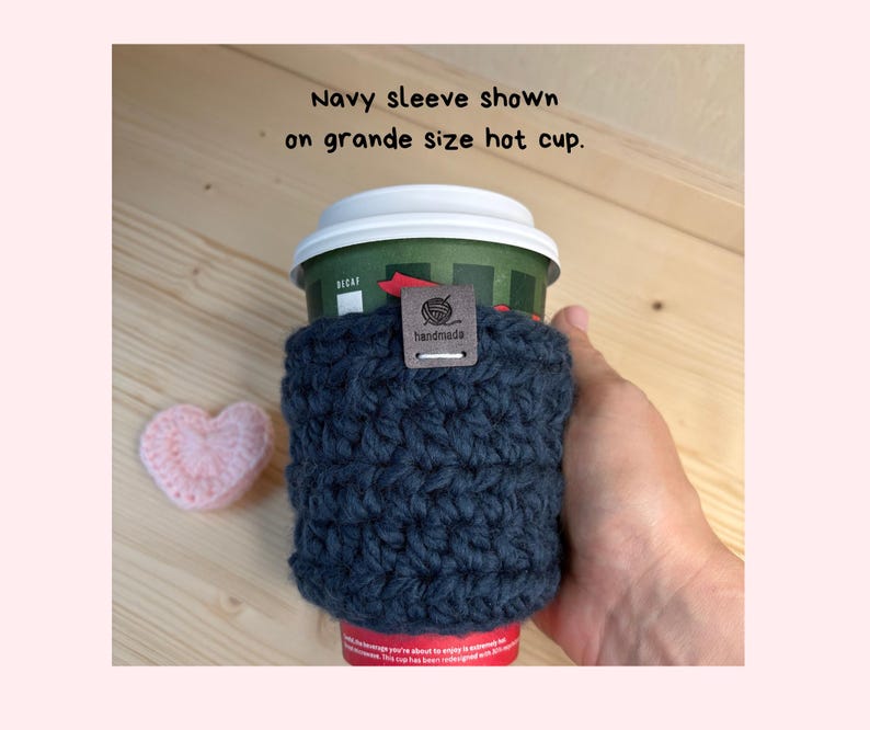 May include: A navy blue crocheted cup sleeve featuring a small brown rectangular tag with the text handmade and a yarn ball icon. The sleeve is wrapped around a green grande size hot cup with a white lid. Text reads: Navy sleeve shown on grande size hot cup.