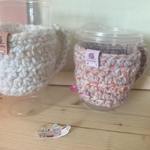 May include: Two clear plastic cups with handles, each featuring a thick, crocheted cosy. One cosy is solid white with subtle metallic flecks, and the other is a variegated blend of pink, orange, and white. Both have a small tag labelled handmade.