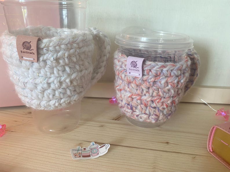 May include: Two clear plastic cups with handles, each featuring a thick, crocheted cosy. One cosy is solid white with subtle metallic flecks, and the other is a variegated blend of pink, orange, and white. Both have a small tag labelled handmade.