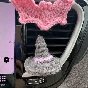 Puede incluir: Un murci&eacute;lago de ganchillo rosa colocado sobre un sombrero de bruja de ganchillo gris con una banda rosa claro. Ambos objetos est&aacute;n sujetos a las lamas horizontales de pl&aacute;stico negro de una rejilla de ventilaci&oacute;n junto a una pantalla.