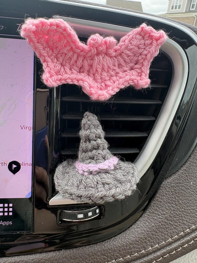 Puede incluir: Un murci&eacute;lago de ganchillo rosa colocado sobre un sombrero de bruja de ganchillo gris con una banda rosa claro. Ambos objetos est&aacute;n sujetos a las lamas horizontales de pl&aacute;stico negro de una rejilla de ventilaci&oacute;n junto a una pantalla.