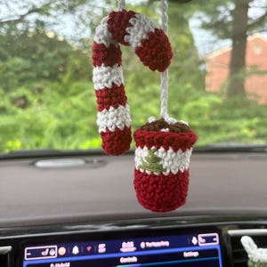 May include: A crocheted hanging ornament featuring a red and white striped candy cane paired with a small red and white mug filled with brown liquid, accented by a green embroidered tree design, all suspended by a white string.