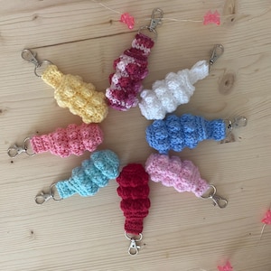 Puede incluir: Una colección de ocho llaveros de ganchillo dispuestos en un patrón circular. Cada llavero presenta un diseño texturizado similar a burbujas en varios colores sólidos, incluyendo amarillo, rosa, blanco, azul, azul claro, rojo y rosa y blanco.