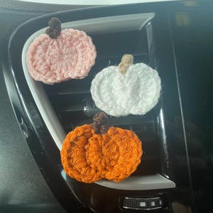Puede incluir: Tres peque&ntilde;os adornos de calabaza tejidos a ganchillo en rosa claro, blanco y naranja. Cada calabaza presenta un dise&ntilde;o texturizado y acanalado con un peque&ntilde;o tallo de hilo en contraste unido a la parte superior. Las calabazas est&aacute;n dispuestas en grupo, mostrando sus colores distintos y su construcci&oacute;n artesanal de hilo.