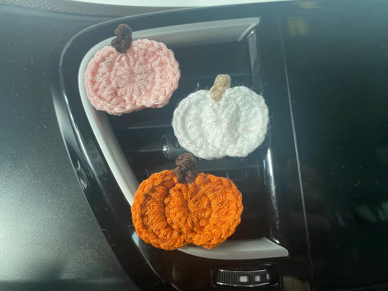 Puede incluir: Tres peque&ntilde;os adornos de calabaza tejidos a ganchillo en rosa claro, blanco y naranja. Cada calabaza presenta un dise&ntilde;o texturizado y acanalado con un peque&ntilde;o tallo de hilo en contraste unido a la parte superior. Las calabazas est&aacute;n dispuestas en grupo, mostrando sus colores distintos y su construcci&oacute;n artesanal de hilo.