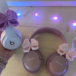 May include: A pair of pink headphones with crocheted bows attached. One set of headphones is white with a purple bow, and the other is pink with two pink bows. The headphones are on a wooden surface.