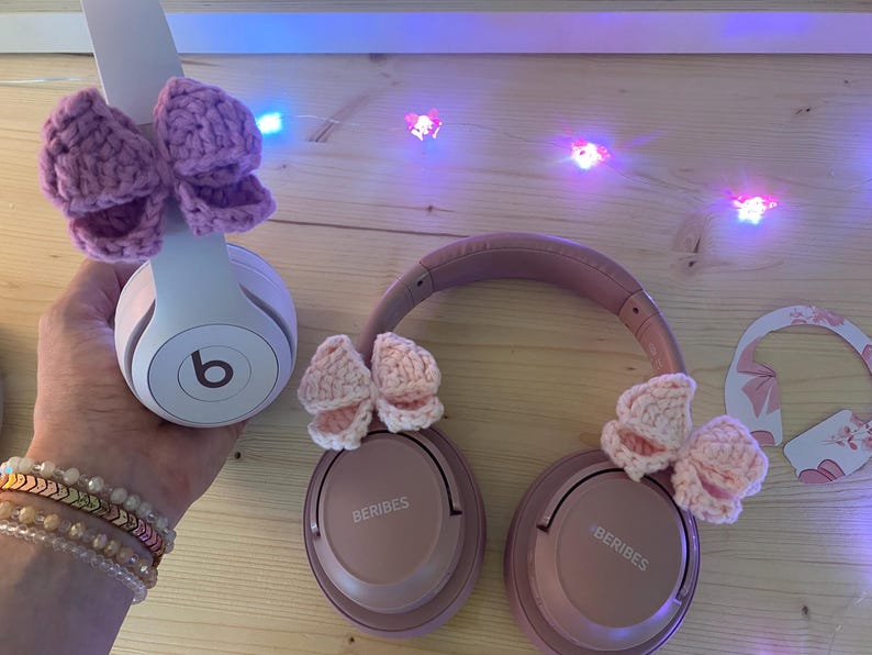 May include: A pair of pink headphones with crocheted bows attached. One set of headphones is white with a purple bow, and the other is pink with two pink bows. The headphones are on a wooden surface.
