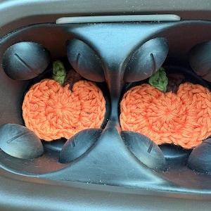 Puede incluir: Dos pequeños posavasos de ganchillo con forma de calabaza naranja, con tallos verdes y detalles marrones, colocados cada uno en un portavasos circular de plástico negro. Las calabazas de hilo texturizado tienen un patrón de punto circular.
