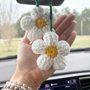 May include: Two white crochet daisy car charms with yellow centers and green hanging strings. The daisy petals are fluffy and textured. The charms are hanging in front of a car dashboard.