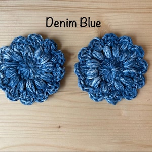 May include: Two round, denim blue crochet coasters featuring a textured, floral-inspired puff stitch pattern. The yarn displays a variegated mix of dark and light blue tones. The text "Denim Blue" is printed in black above the two circular items.