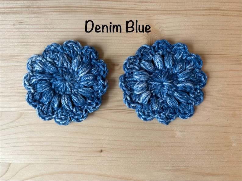 May include: Two round, denim blue crochet coasters featuring a textured, floral-inspired puff stitch pattern. The yarn displays a variegated mix of dark and light blue tones. The text "Denim Blue" is printed in black above the two circular items.