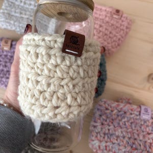 May include: A clear glass tumbler with a bamboo lid, featuring a thick, cream-coloured crocheted sleeve. A small, rectangular brown leather tag is attached to the sleeve, embossed with a yarn ball icon and the word "handmade" in white text.