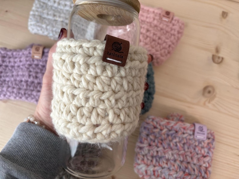 May include: A clear glass tumbler with a bamboo lid, featuring a thick, cream-coloured crocheted sleeve. A small, rectangular brown leather tag is attached to the sleeve, embossed with a yarn ball icon and the word "handmade" in white text.
