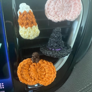 May include: Crocheted Halloween-themed car air fresheners. The set includes a candy corn, a pink pumpkin, an orange pumpkin, and a black witch hat with a purple band. These handmade decorations add a festive touch to any vehicle.