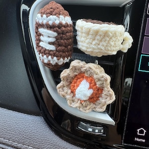 Puede incluir: Tres peque&ntilde;os adornos de ganchillo sujetos a la rejilla de ventilaci&oacute;n de un coche. Uno es un bal&oacute;n de f&uacute;tbol americano marr&oacute;n y blanco, otro es una taza de caf&eacute; color crema y otro es una pieza en forma de flor con borde beige, centro naranja y blanco.