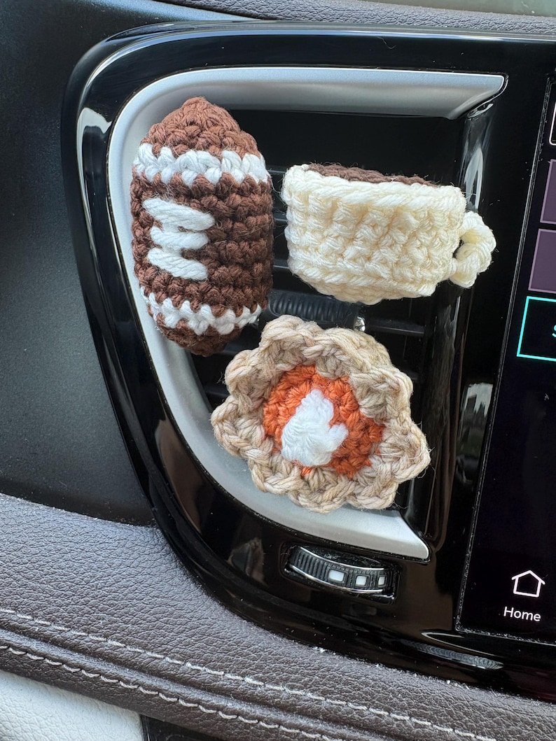 Puede incluir: Tres peque&ntilde;os adornos de ganchillo sujetos a la rejilla de ventilaci&oacute;n de un coche. Uno es un bal&oacute;n de f&uacute;tbol americano marr&oacute;n y blanco, otro es una taza de caf&eacute; color crema y otro es una pieza en forma de flor con borde beige, centro naranja y blanco.