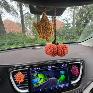 Puede incluir: Un conjunto de accesorios de coche de ganchillo que incluye una hoja amarilla mostaza y una calabaza naranja quemado colgando del espejo retrovisor, junto a dos hojas de ganchillo m&aacute;s peque&ntilde;as en naranja y magenta en las rejillas.