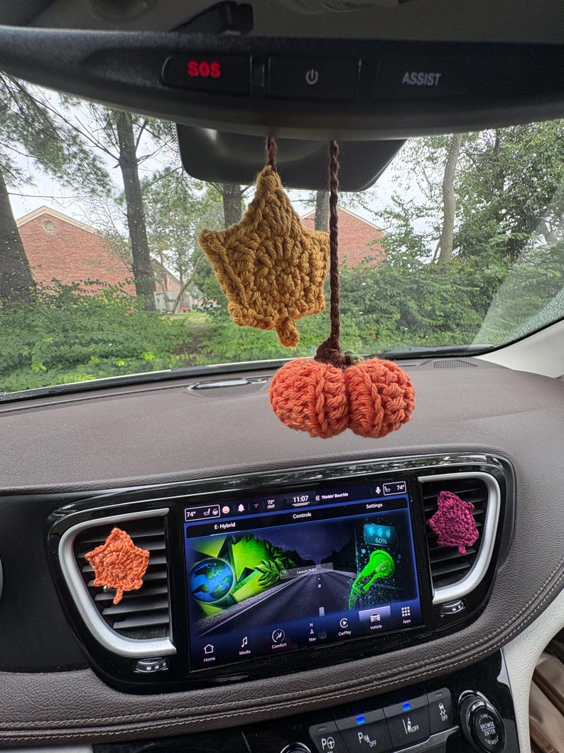 Puede incluir: Un conjunto de accesorios de coche de ganchillo que incluye una hoja amarilla mostaza y una calabaza naranja quemado colgando del espejo retrovisor, junto a dos hojas de ganchillo m&aacute;s peque&ntilde;as en naranja y magenta en las rejillas.