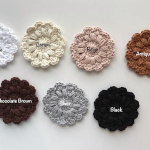 May include: Seven circular crochet flower appliques arranged in two rows. Colors include White Sparkle, Cream, Beige, Antique Gold, Chocolate Brown, Silver, and Black. Each piece features a textured, puff-stitch petal design with a central label.