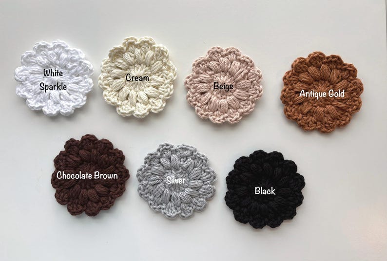 May include: Seven circular crochet flower appliques arranged in two rows. Colors include White Sparkle, Cream, Beige, Antique Gold, Chocolate Brown, Silver, and Black. Each piece features a textured, puff-stitch petal design with a central label.
