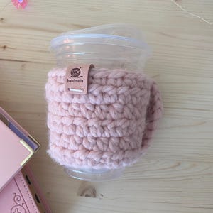 May include: A light pink crochet cup sleeve wrapped around a clear plastic cup with a lid. A small tan leather tag is attached to the top edge of the sleeve, featuring a graphic of a ball of yarn and the text "handmade" printed in black ink.