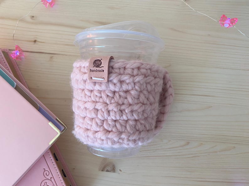 May include: A light pink crochet cup sleeve wrapped around a clear plastic cup with a lid. A small tan leather tag is attached to the top edge of the sleeve, featuring a graphic of a ball of yarn and the text "handmade" printed in black ink.