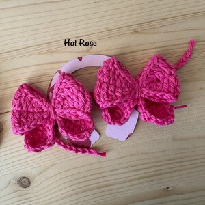 May include: Two hot pink crochet bows with a horseshoe-shaped cutout. The bows are made of a textured yarn and have a detailed, handmade appearance. The text "Hot Rose" is above the bows. The background is a light wood surface.