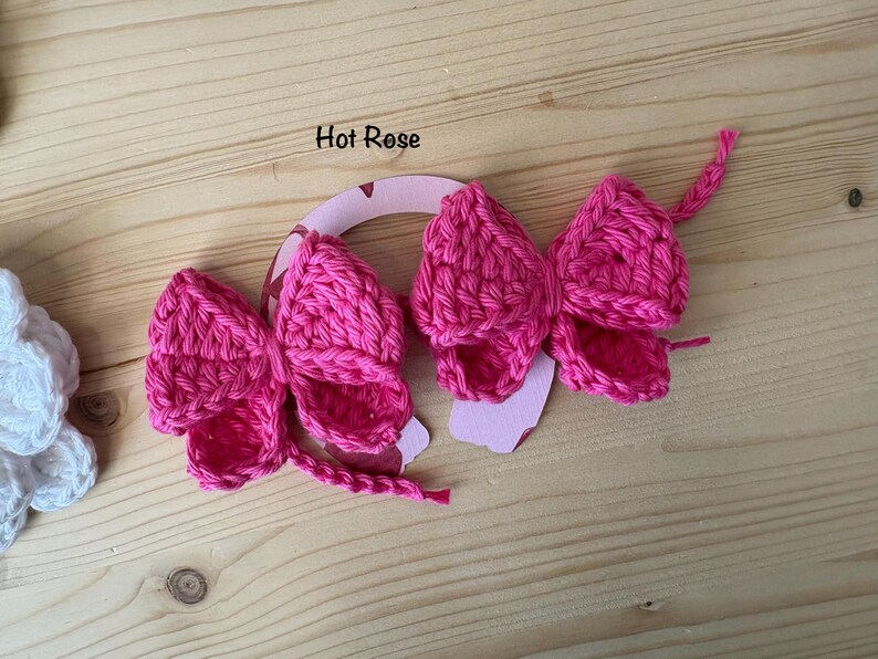 May include: Two hot pink crochet bows with a horseshoe-shaped cutout. The bows are made of a textured yarn and have a detailed, handmade appearance. The text "Hot Rose" is above the bows. The background is a light wood surface.