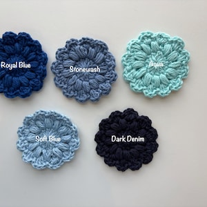 May include: Five circular crochet flower appliques in various shades of blue. The colors are labeled as Royal Blue, Stonewash, Aqua, Soft Blue, and Dark Denim. Each piece features a textured, puff-stitch petal design arranged in a radial pattern.