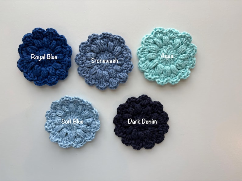 May include: Five circular crochet flower appliques in various shades of blue. The colors are labeled as Royal Blue, Stonewash, Aqua, Soft Blue, and Dark Denim. Each piece features a textured, puff-stitch petal design arranged in a radial pattern.