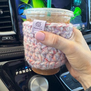 May include: A clear plastic iced coffee cup with a green straw, encased in a thick, hand-crocheted sleeve featuring a variegated yarn pattern of soft pink, purple, and white. A small, rectangular light purple tag is attached, reading "handmade".