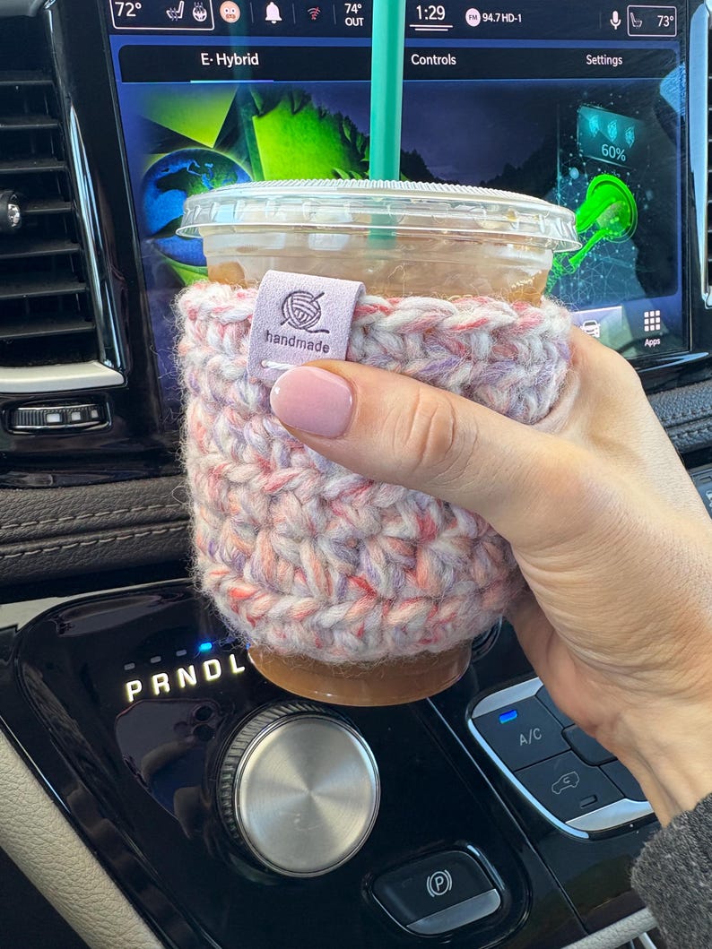 May include: A clear plastic iced coffee cup with a green straw, encased in a thick, hand-crocheted sleeve featuring a variegated yarn pattern of soft pink, purple, and white. A small, rectangular light purple tag is attached, reading "handmade".