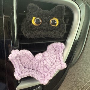 Puede incluir: Una cabeza de gato tejida a ganchillo en color negro con grandes ojos de pl&aacute;stico con borde amarillo y una forma de murci&eacute;lago tejida en color lila, dise&ntilde;ados como clips decorativos para ventilaci&oacute;n. El gato tiene orejas puntiagudas.