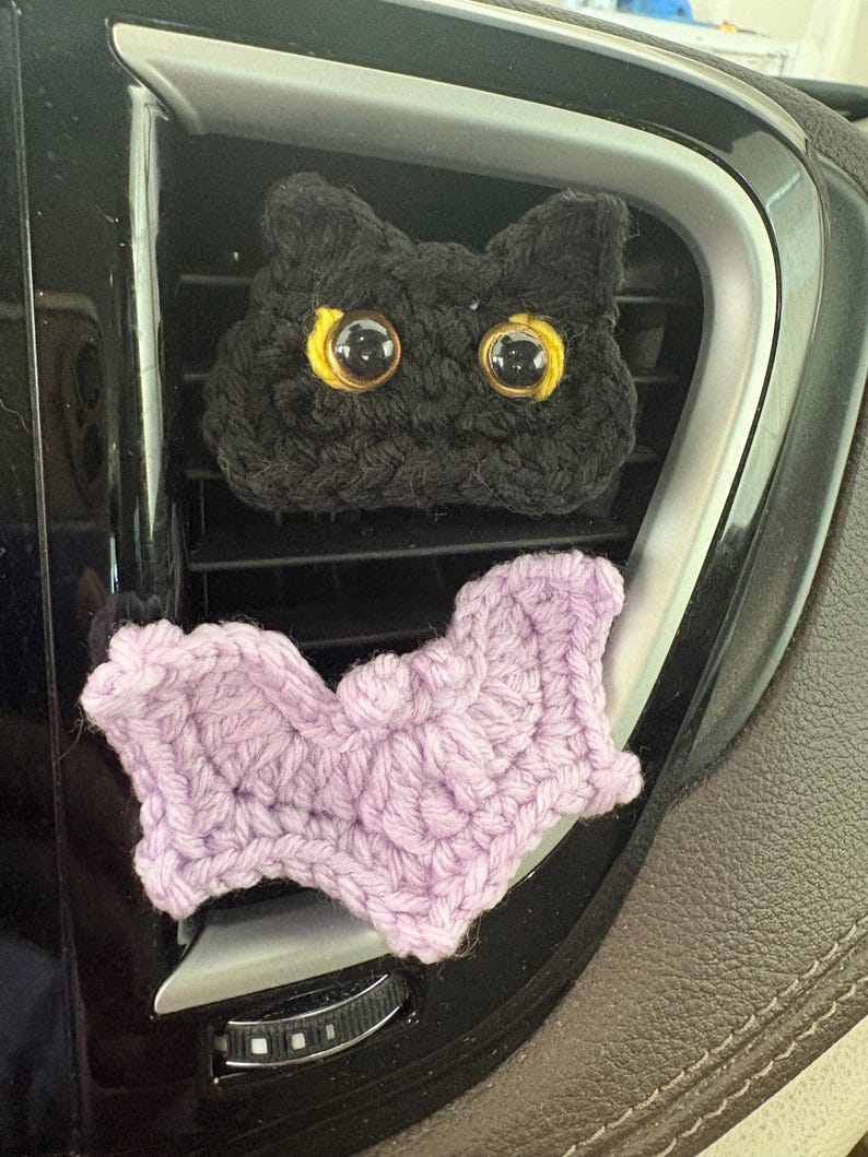 Puede incluir: Una cabeza de gato tejida a ganchillo en color negro con grandes ojos de pl&aacute;stico con borde amarillo y una forma de murci&eacute;lago tejida en color lila, dise&ntilde;ados como clips decorativos para ventilaci&oacute;n. El gato tiene orejas puntiagudas.