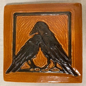May include: A square ceramic tile featuring a relief design of two dark brown ravens facing each other. The background of the central square is a lighter amber-orange color with a textured finish, framed by a dark brown border on a glossy orange tile.