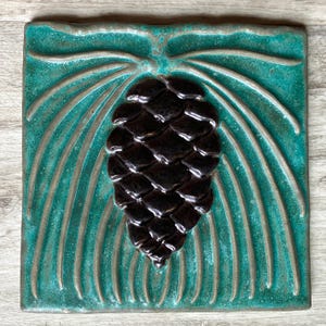 May include: A square ceramic tile featuring a raised, dark brown pinecone relief centered against a textured, vibrant teal background. The design includes embossed, sweeping pine needle patterns that radiate outward from the top of the pinecone.