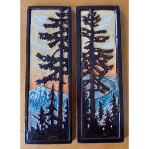 May include: Two rectangular ceramic wall art pieces. Each features a dark brown tree silhouette against a landscape of blue mountains, orange, and yellow sky. The pieces have a dark brown border.