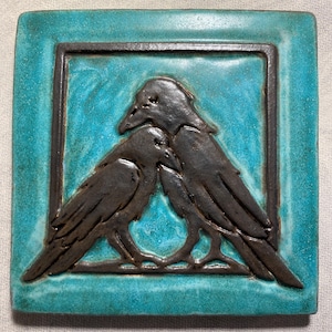 May include: A square ceramic tile featuring a relief design of two dark brown ravens facing each other. The background of the tile is a vibrant turquoise blue with a glossy finish, framed by a dark brown border that matches the color of the birds.