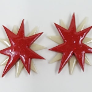 Pu&ograve; includere: Un paio di orecchini a forma di stella rossa e bianca. Le stelle sono sovrapposte, con una stella rossa pi&ugrave; piccola sopra una stella bianca pi&ugrave; grande.