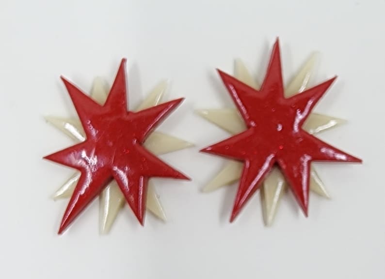 Pu&ograve; includere: Un paio di orecchini a forma di stella rossa e bianca. Le stelle sono sovrapposte, con una stella rossa pi&ugrave; piccola sopra una stella bianca pi&ugrave; grande.