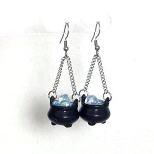 May include: A pair of dangle earrings featuring miniature black cauldrons suspended from silver-toned metal chains. Each cauldron contains small, iridescent, light blue spherical accents that resemble bubbling liquid, attached to standard silver-toned ear hooks.