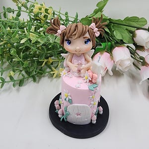 May include: A decorative figurine featuring a small character with brown pigtails and blue eyes sitting atop a cylindrical pink base. The base is adorned with white daisy flowers, green leaves, and a small white door. The entire piece sits on a black circular stand.