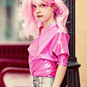 May include: A person wearing a bright pink metallic short-sleeved top and a silver-toned waist belt with long fringe detailing. The outfit is paired with a vibrant pink wig, matching pink eye makeup, and star-shaped earrings in a matching shade.
