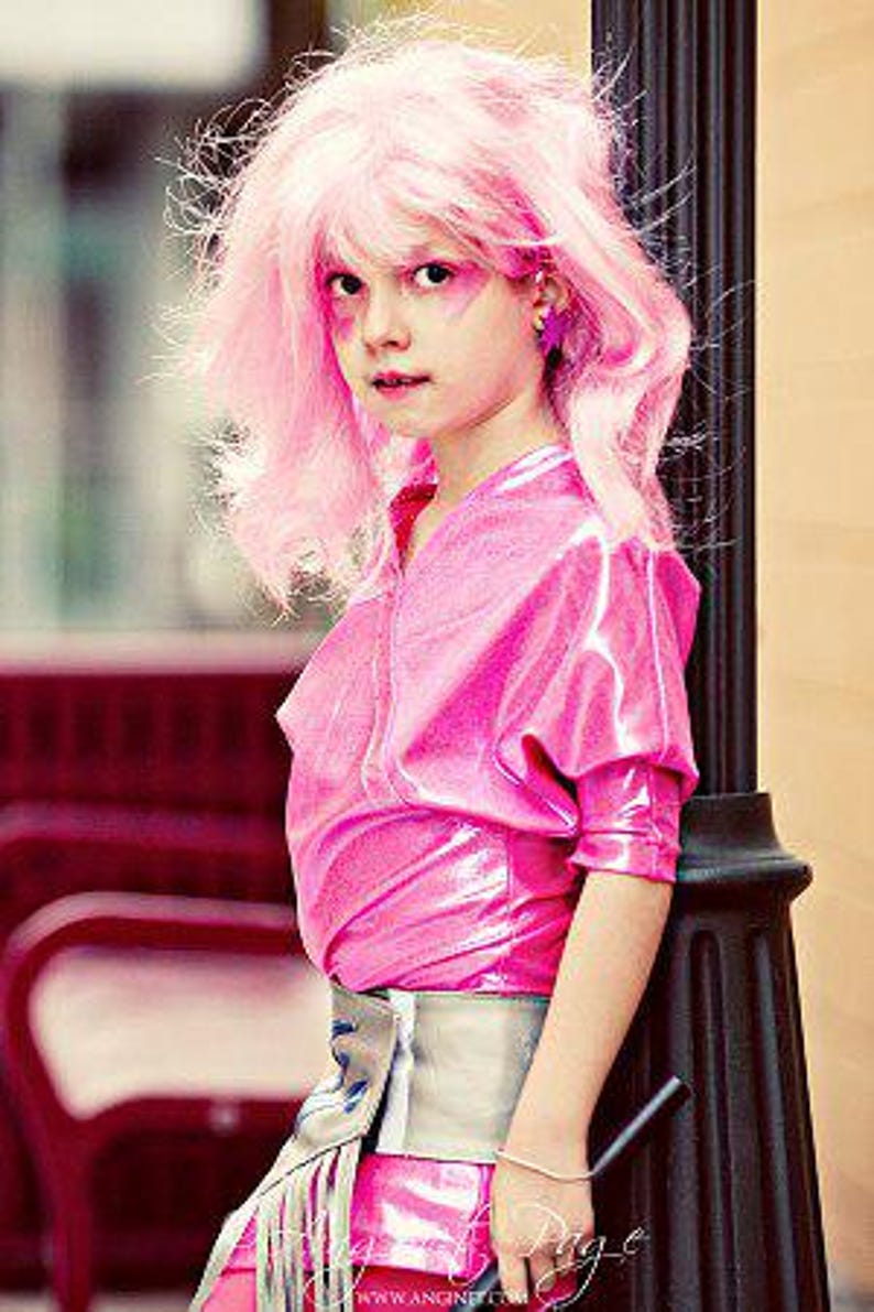 May include: A person wearing a bright pink metallic short-sleeved top and a silver-toned waist belt with long fringe detailing. The outfit is paired with a vibrant pink wig, matching pink eye makeup, and star-shaped earrings in a matching shade.