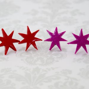 May include: Four star-shaped stud earrings arranged in a row. The two earrings on the left are a solid, glossy red color, while the two earrings on the right are a solid, glossy magenta color. Each star features eight sharp, pointed tips.
