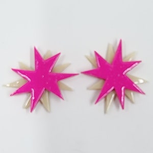 Pu&ograve; includere: Un paio di orecchini a forma di stella rosa e crema. Gli orecchini sono sovrapposti, con una stella rosa pi&ugrave; piccola sopra una stella crema pi&ugrave; grande.