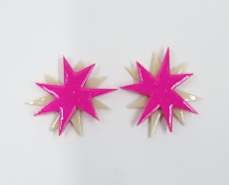 Pu&ograve; includere: Un paio di orecchini a forma di stella rosa e crema. Gli orecchini sono sovrapposti, con una stella rosa pi&ugrave; piccola sopra una stella crema pi&ugrave; grande.