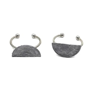 May include: Two silver-toned metal rings, each featuring a semi-circular, dark gray stone. The rings have a simple, open design with a small silver ball at each end of the band. The stone has a textured surface.